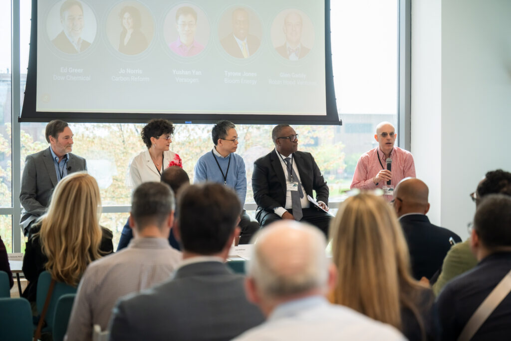 A panel of five speakers in front of an audience. A man in a pink shirt has the mircophone.