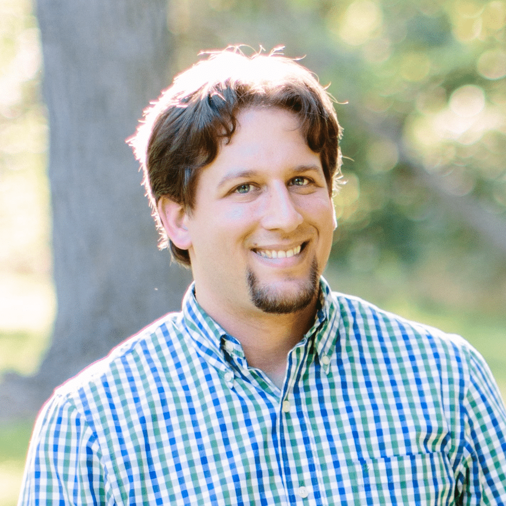 Micheal outside in front of a tree, he has a goatee and a blue and white checkered button down on.