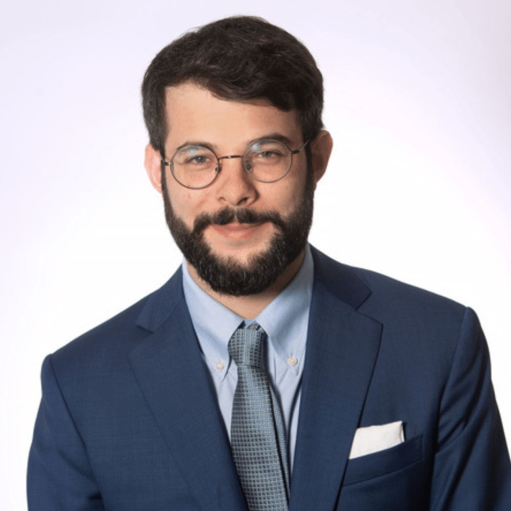 Anthony has dark hair and a mustache and beard. He is wearing a navy-blue suit jacket, a light blue collared shirt and a grey tie.