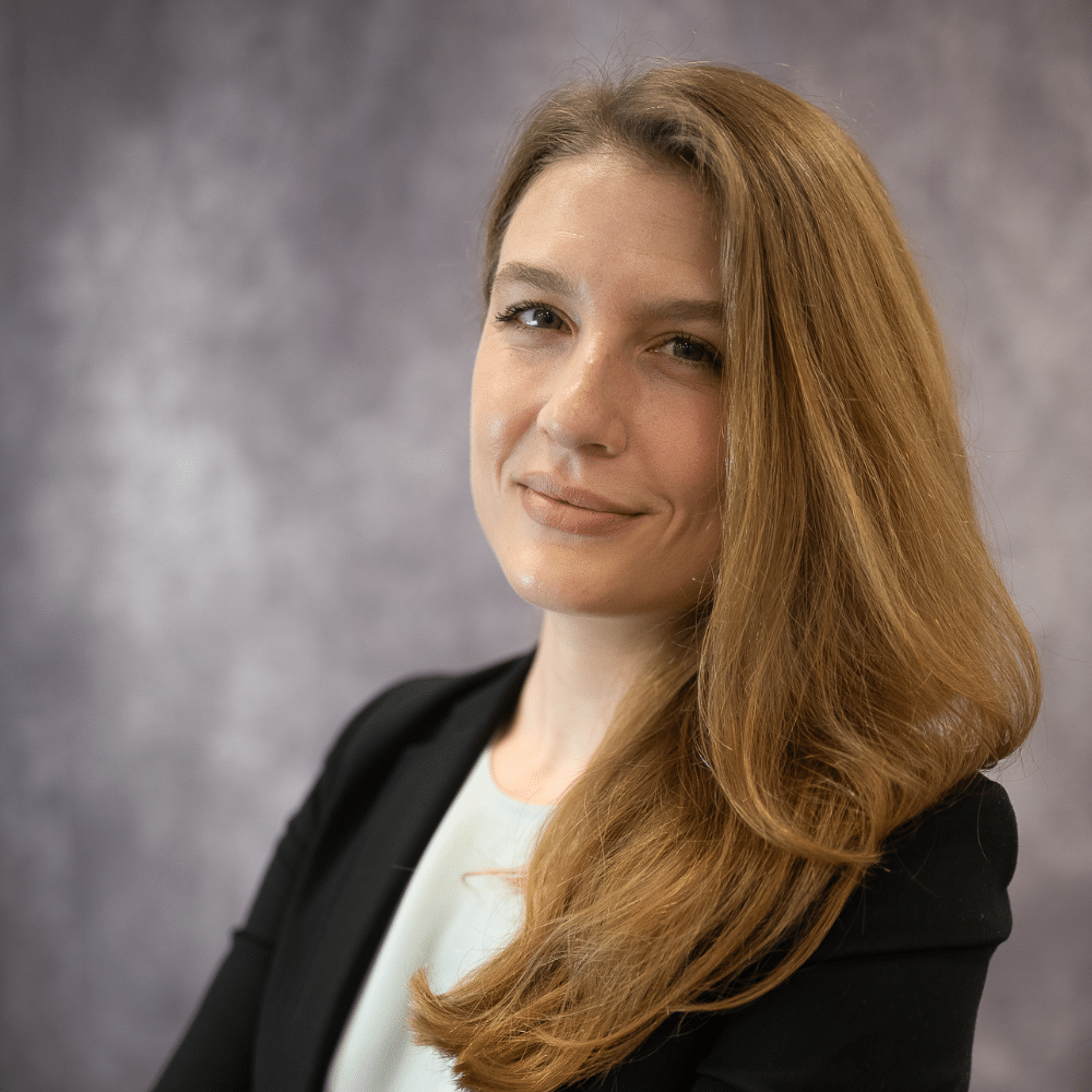 Katie is standing in front of a grey wall with her arms crossed. She has a black dress jacket on and a lighter top underneath.