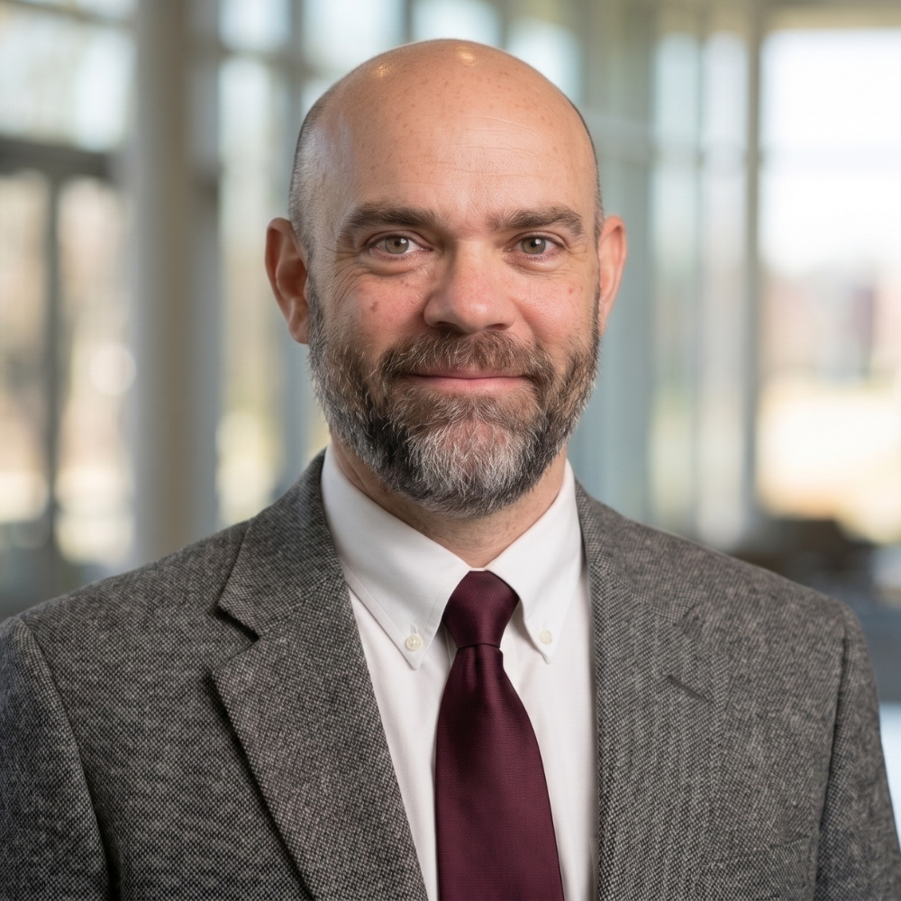 Paul is standing in a room full of windows, wearing a tweed-like suit jacket and maroon tie.