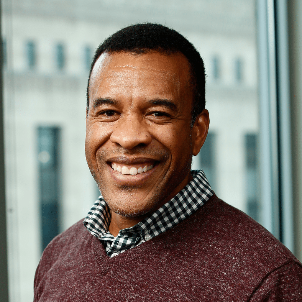 Troy is standing in front of a window with a light blue curtain. He is wearing a burgundy sweater with a checked button down underneath.