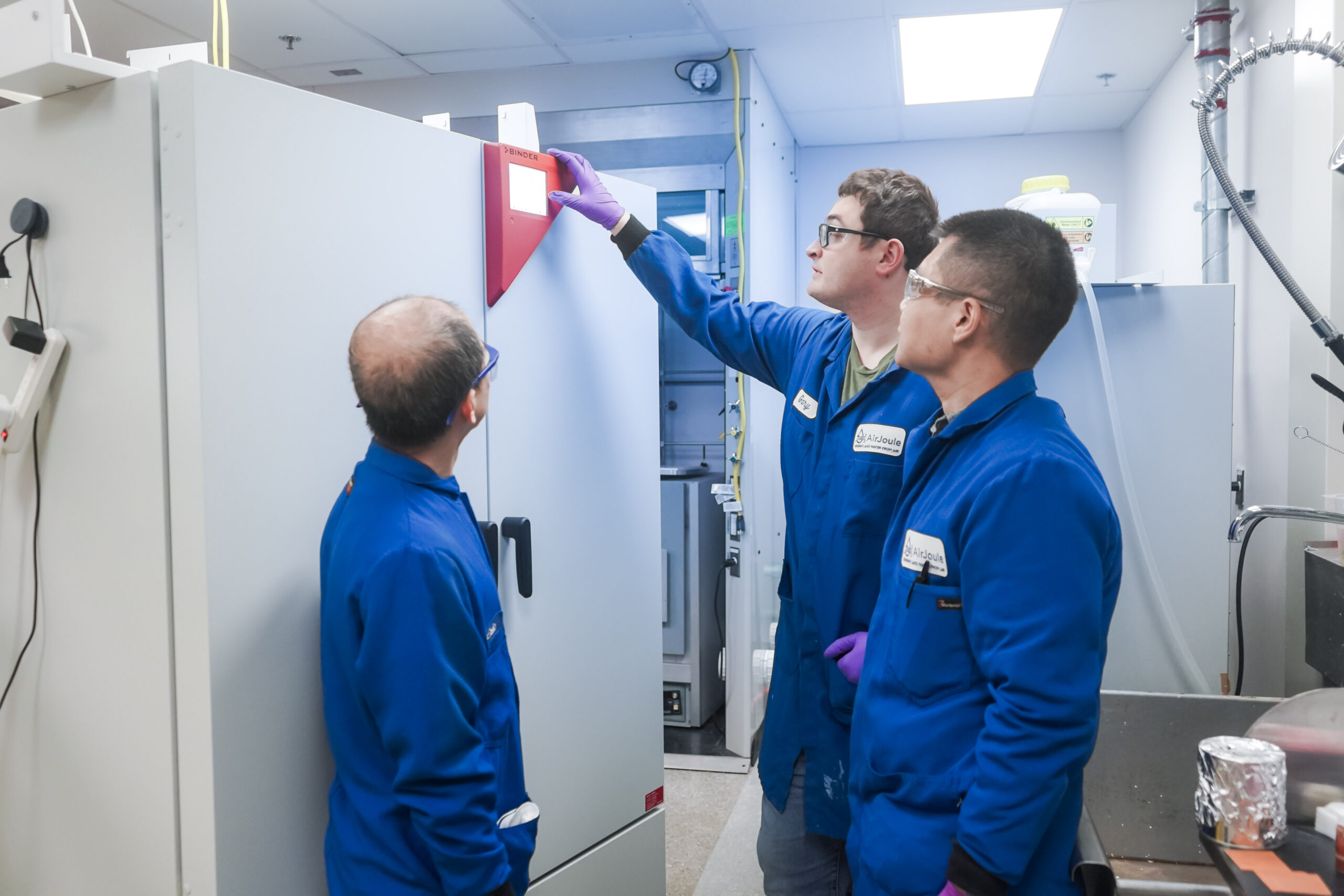 Three men wearing blue lab coats in the AirJoule lab look at a meter.