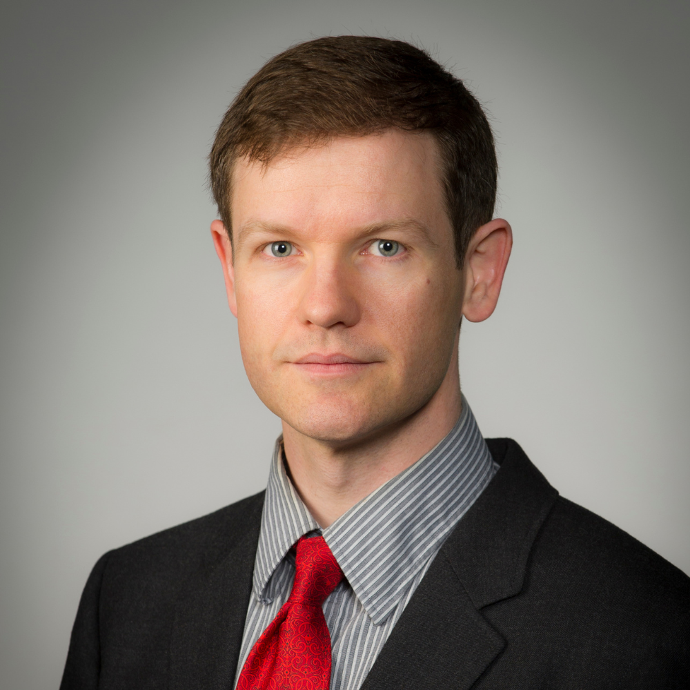Jeff Wilbur is wearing a black suit jacket, a grey and white striped button shirt underneath and a red tie. He is in front of a grey wall.