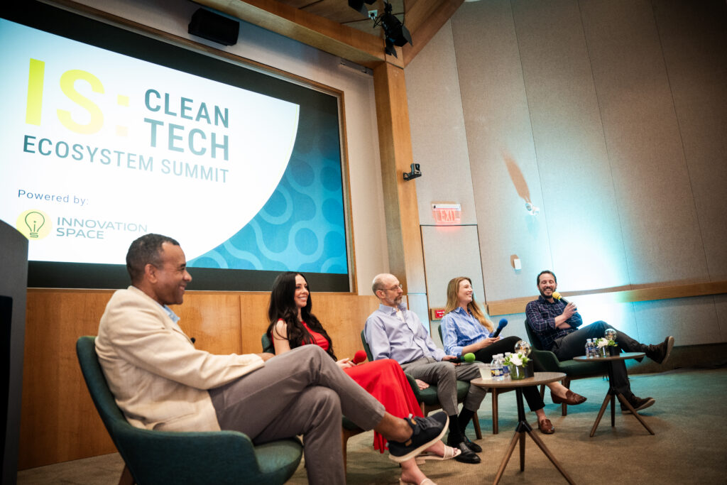 A panel of three men and two ladies sit on a stage with microphones, in front of a large screen that says IS:CleanTech Ecosystem Summit