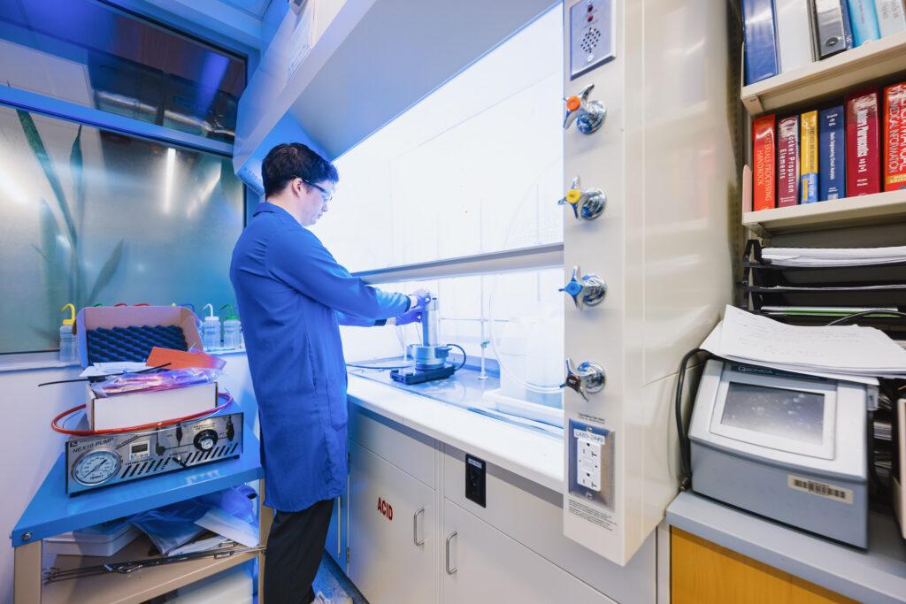 A dark-haired man in a blue lab coat works at the counter in a lab pod.